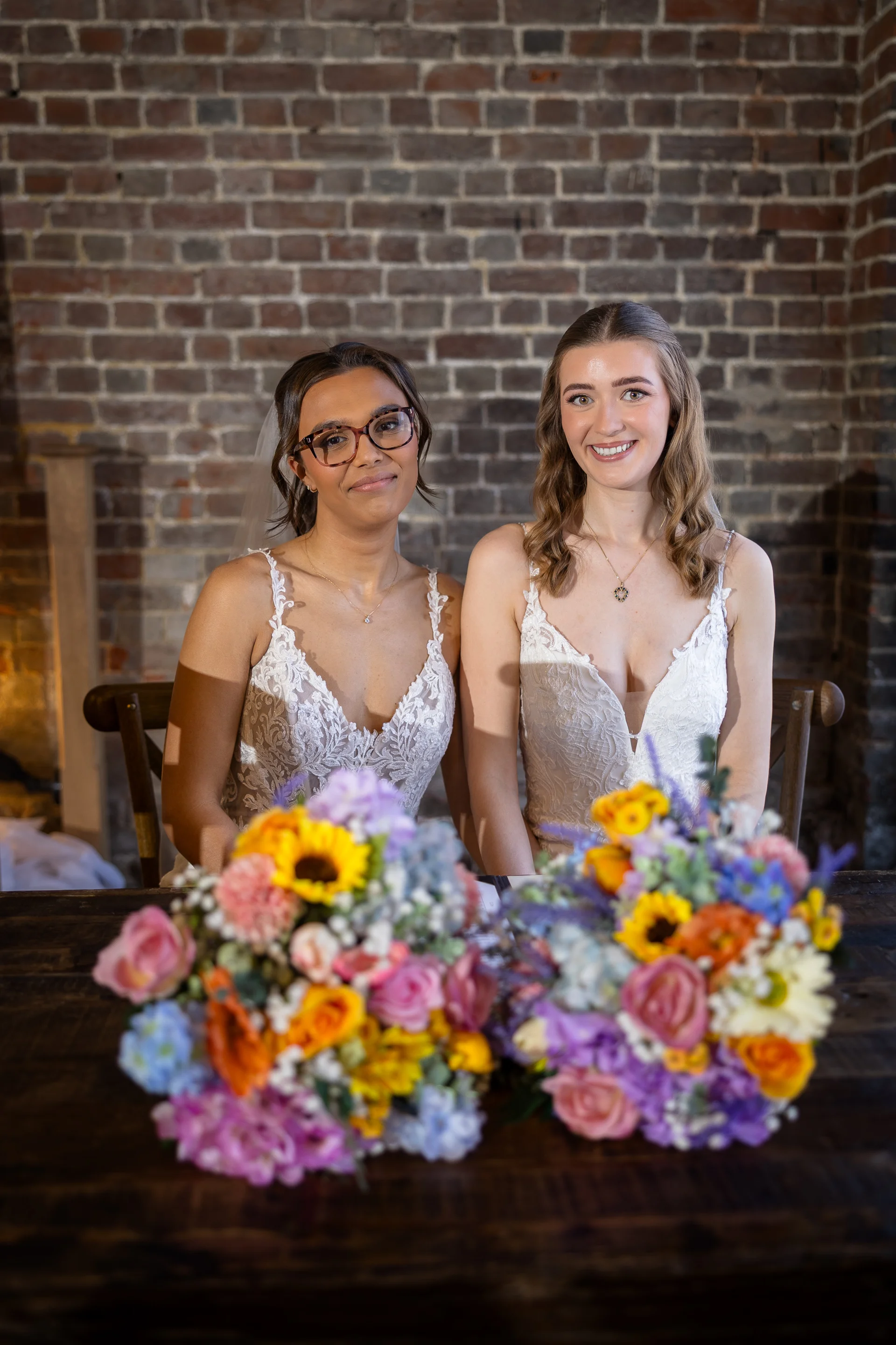 Two smiling brides in matching lace wedding dresses sit together behind colourful floral bouquets against an exposed brick wall.