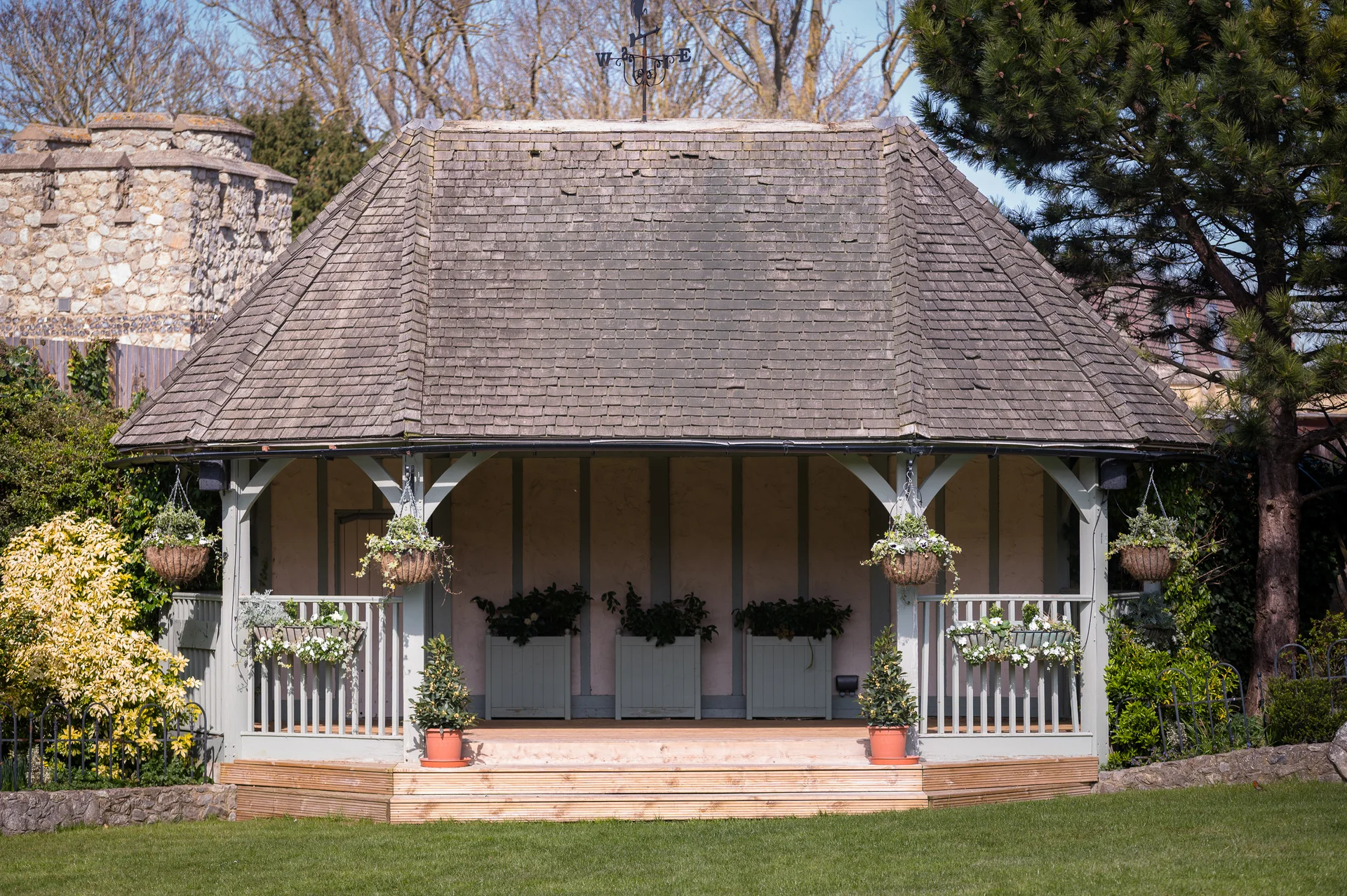 An ornate wooden bandstand with a tiled roof stands on manicured lawns surrounded by lush greenery and historic stone architecture on a sunny day.
