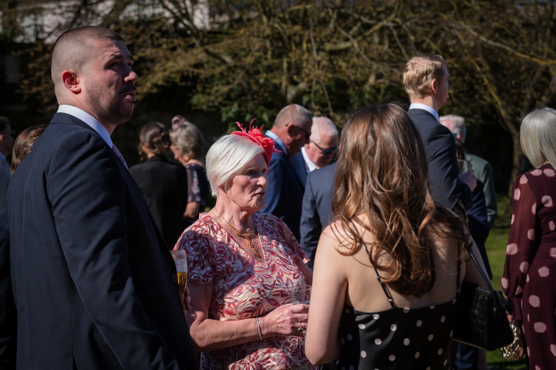Wedding guests mingle and converse outside in the bright sunshine, with a mature woman wearing a patterned top and red fascinator talking to a guest.