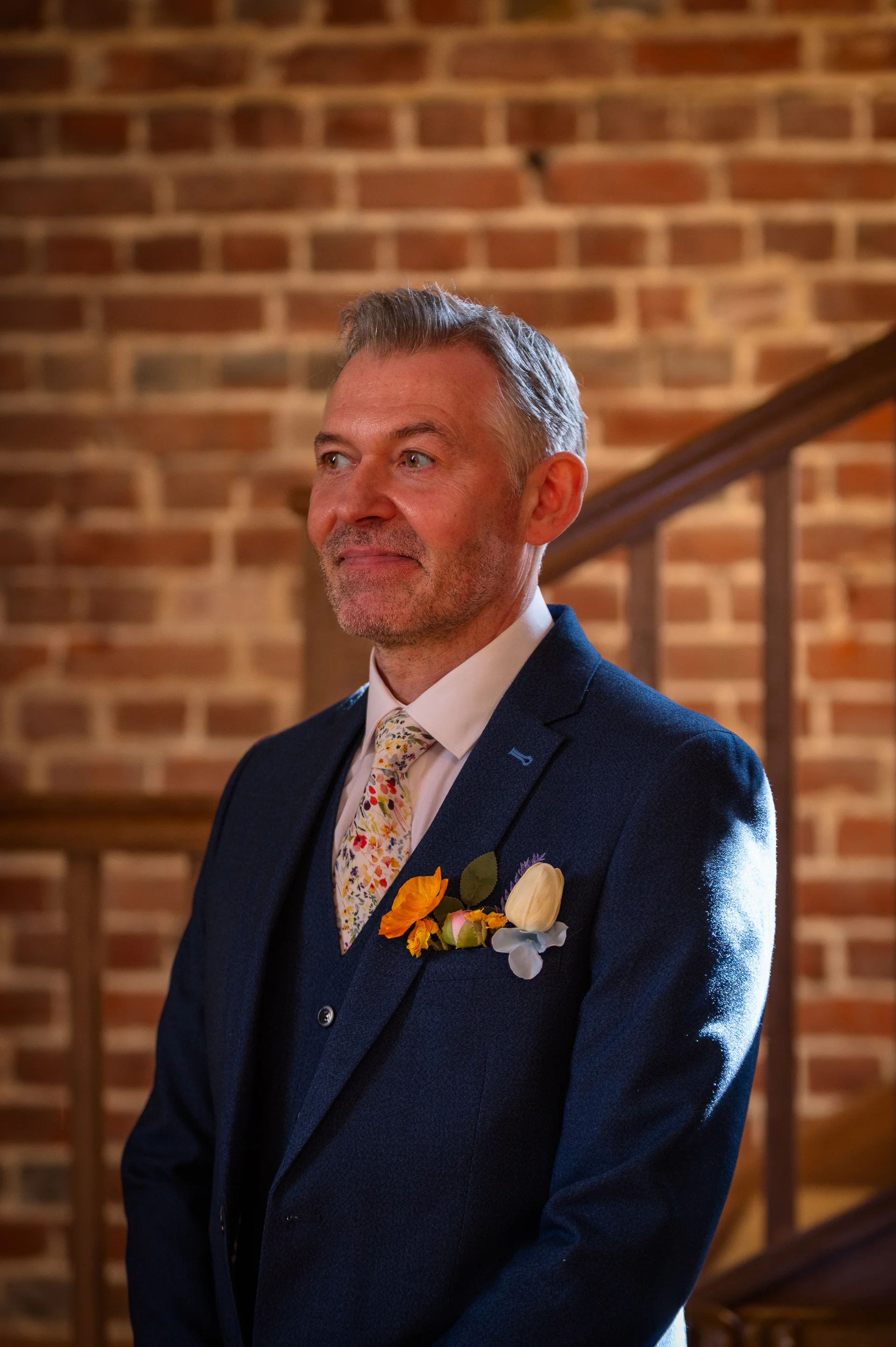 A smiling groom wearing a blue three-piece suit, floral tie, and colourful pocket square stands before an exposed brick wall during his wedding ceremony.
