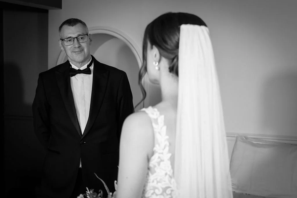 An emotional father in a black tuxedo looks proudly at his daughter wearing a lace wedding dress and veil during a first look moment.