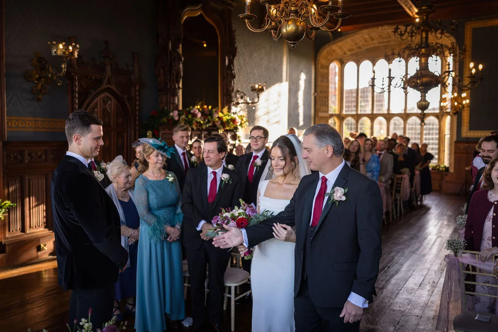 The father of the bride proudly leads her down the aisle to meet the waiting groom in a grand, sunlit historic hall.