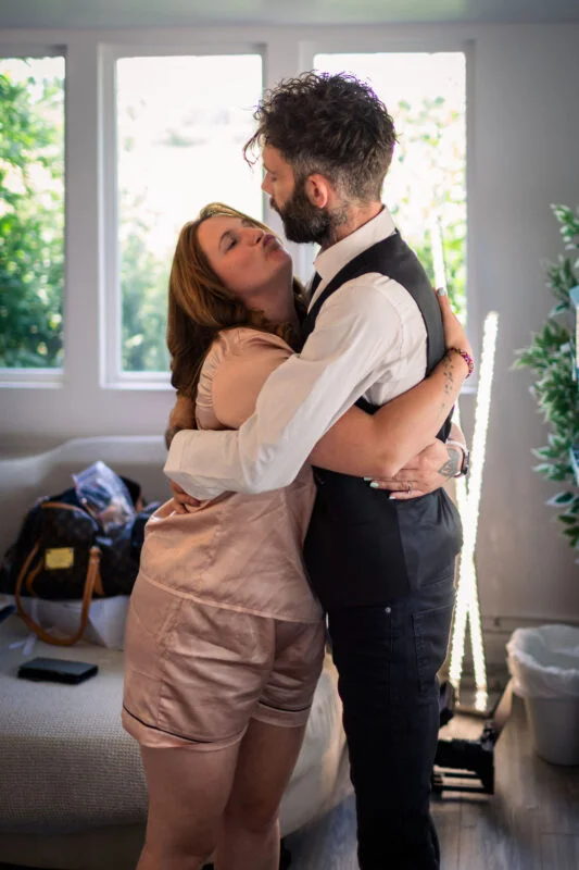 A bride in satin pyjamas and a groom in a waistcoat share a joyful embrace near a bright window during wedding morning preparations.