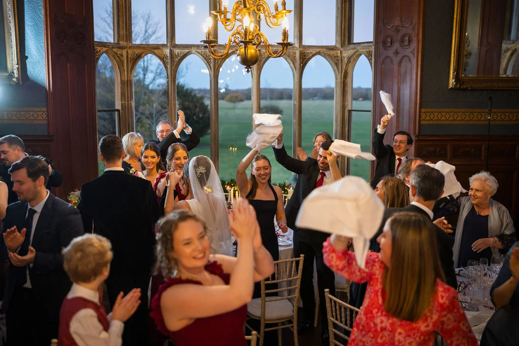 Wedding guests cheer and wave white napkins in the air during speeches inside a grand panelled reception room with large arched windows.