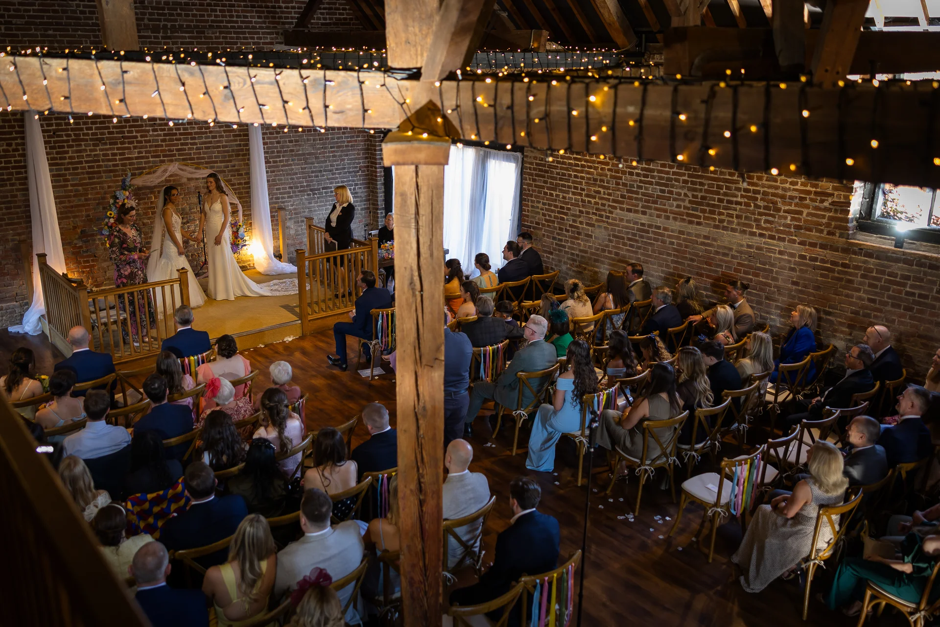 A wide angle shot captures a same-sex wedding ceremony inside a rustic stone and brick barn adorned with a fairy light canopy, featuring two brides standing at the altar with seated guests facing them.