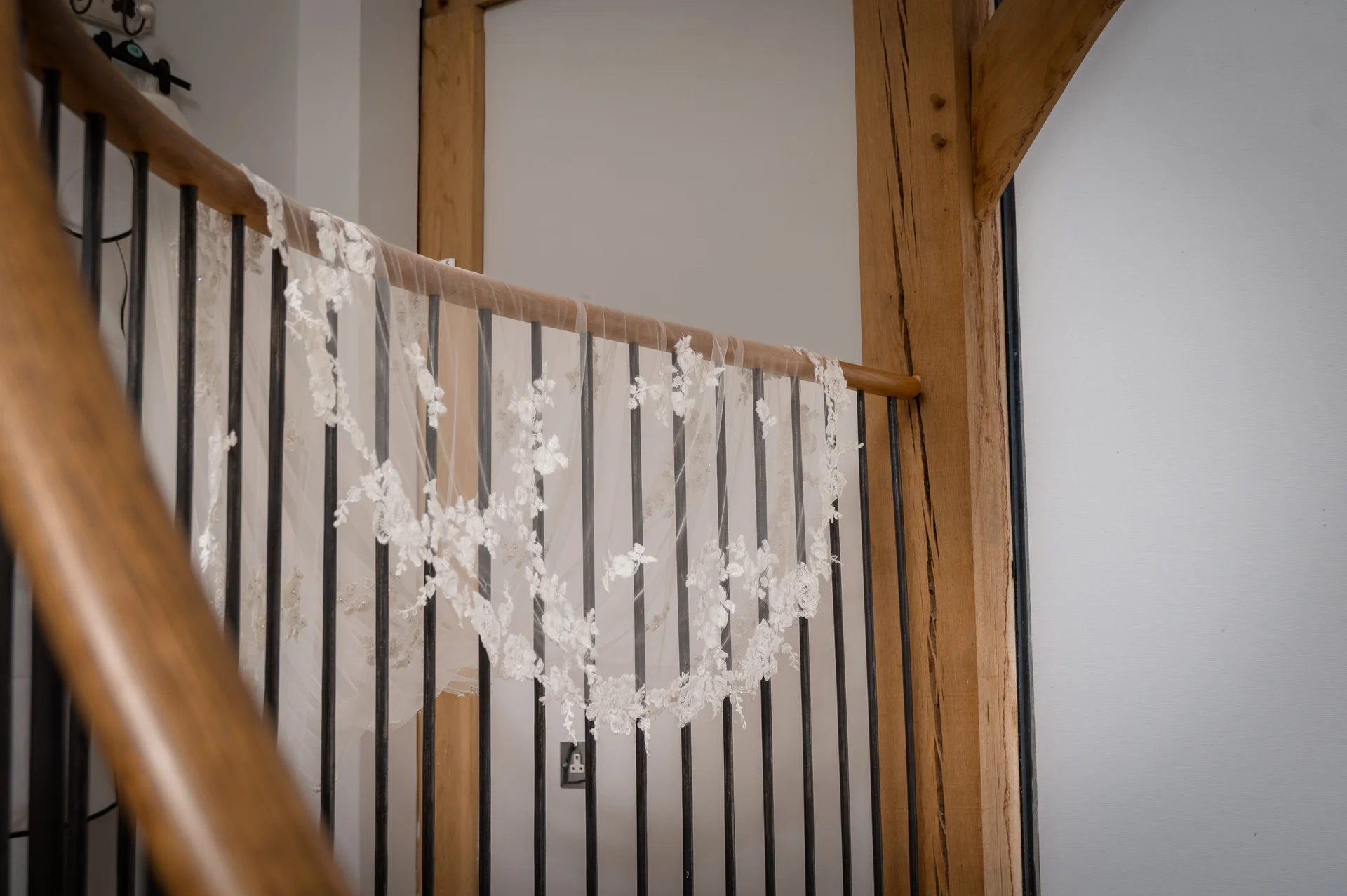 An intricately detailed lace-edged wedding veil is draped elegantly over the wooden banister of a staircase inside a rustic oak-framed barn venue.