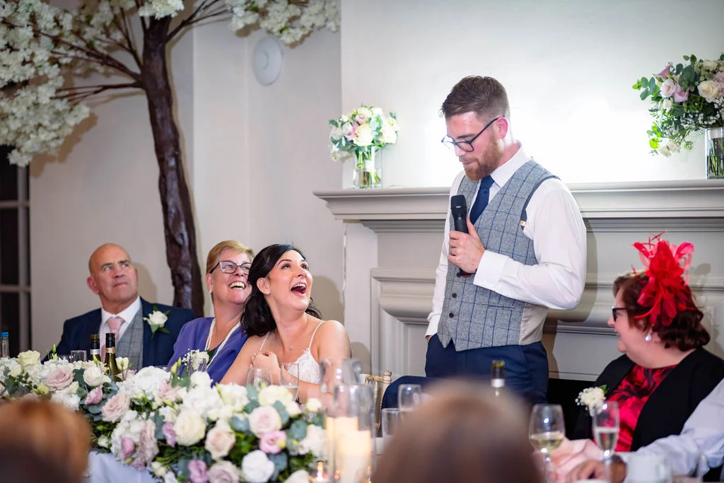 A groom delivers a speech during the wedding reception while his bride and seated guests laugh joyfully at the top table.