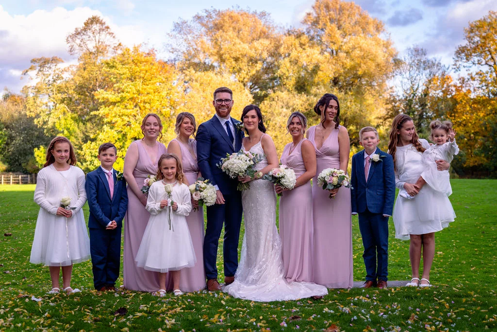 The bride and groom pose outdoors on a green lawn with their wedding party, surrounded by stunning autumn trees with vibrant golden foliage.