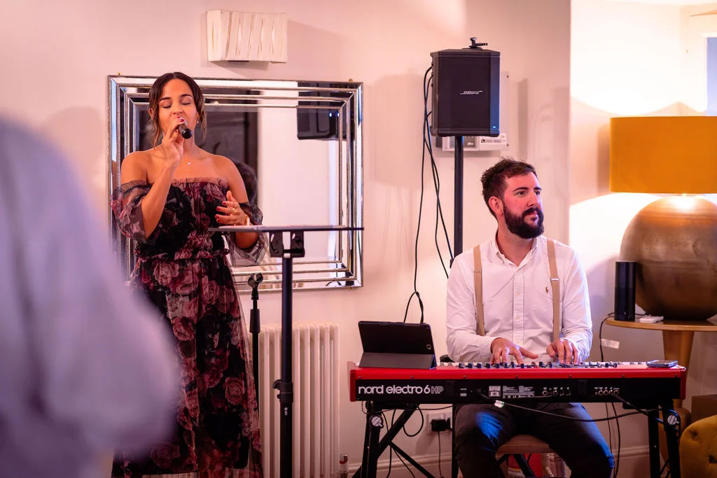 A female vocalist singing into a microphone next to a male keyboard player wearing a white shirt and braces during a wedding reception.