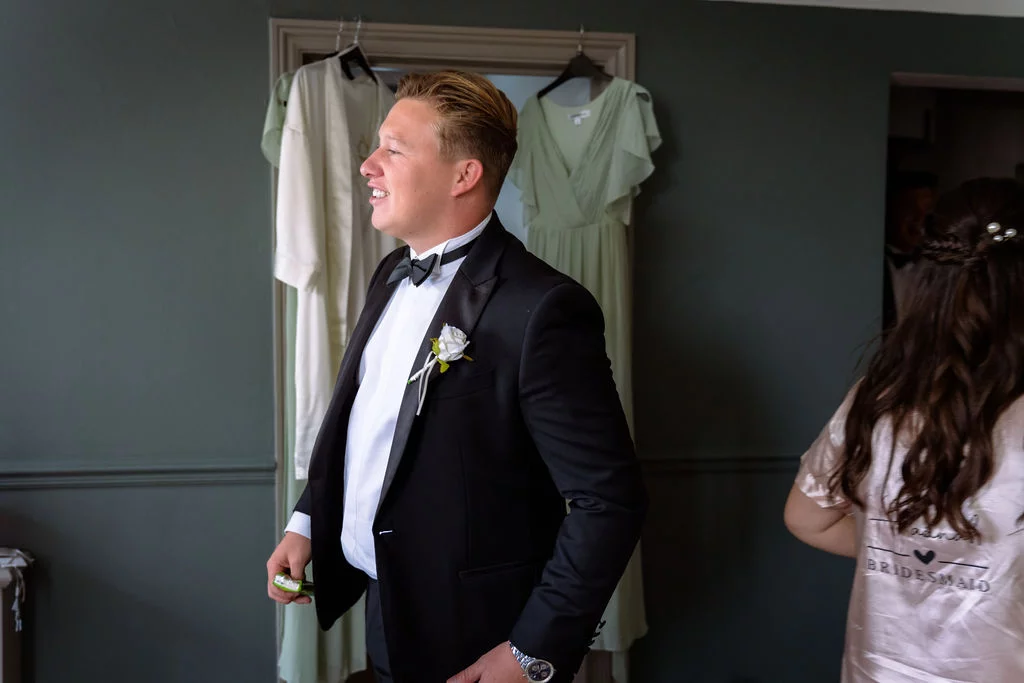 A smiling groom in a black tuxedo and bow tie stands indoors against a dark green wall during wedding morning preparations.