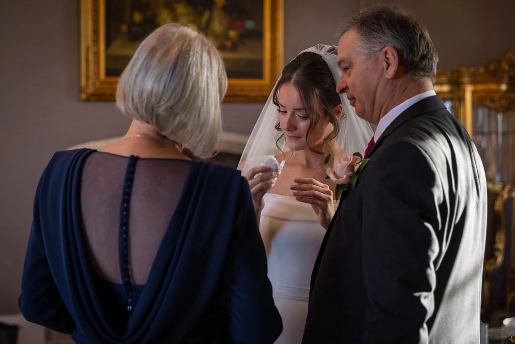 A bride shares an emotional, tearful moment with her parents indoors, surrounded by natural light inside a classic venue.