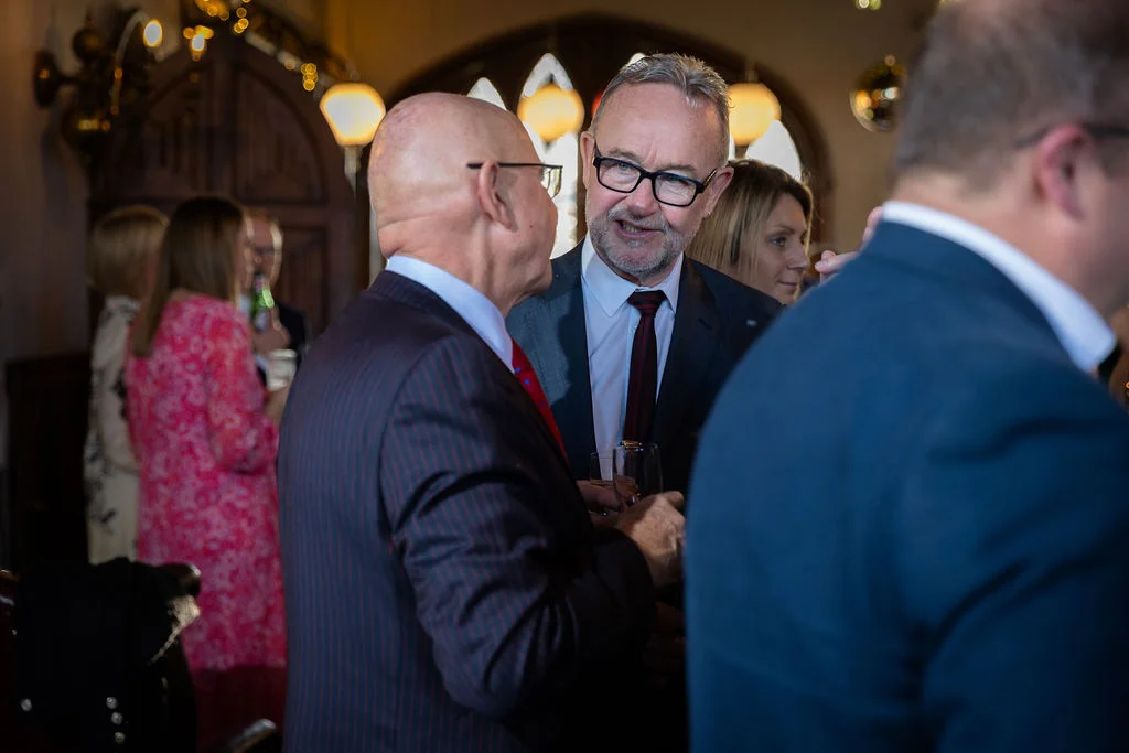 Wedding guests mingling and chatting in a warmly lit venue during the drinks reception, featuring men in tailored suits conversing.