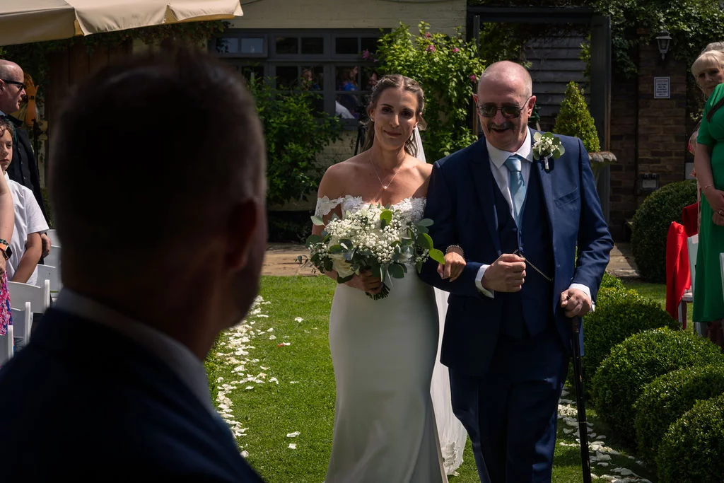 A bride wearing an off-the-shoulder crepe gown and holding a bouquet walks down an outdoor garden aisle on the arm of her father, as the groom looks on from the foreground.