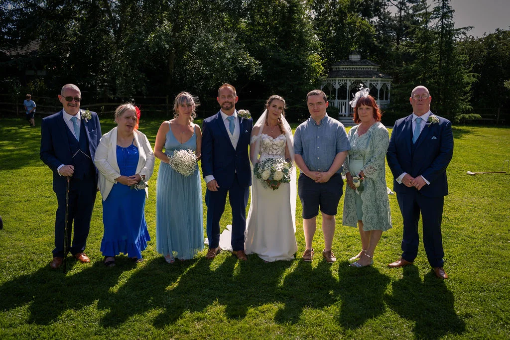 A formal wedding group portrait stands on a sunlit lawn outdoors with a white gazebo in the background.
