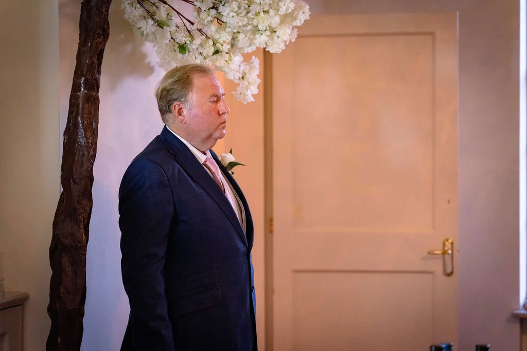 A groom in a navy suit stands waiting at the altar next to an artificial cherry blossom tree inside a venue.
