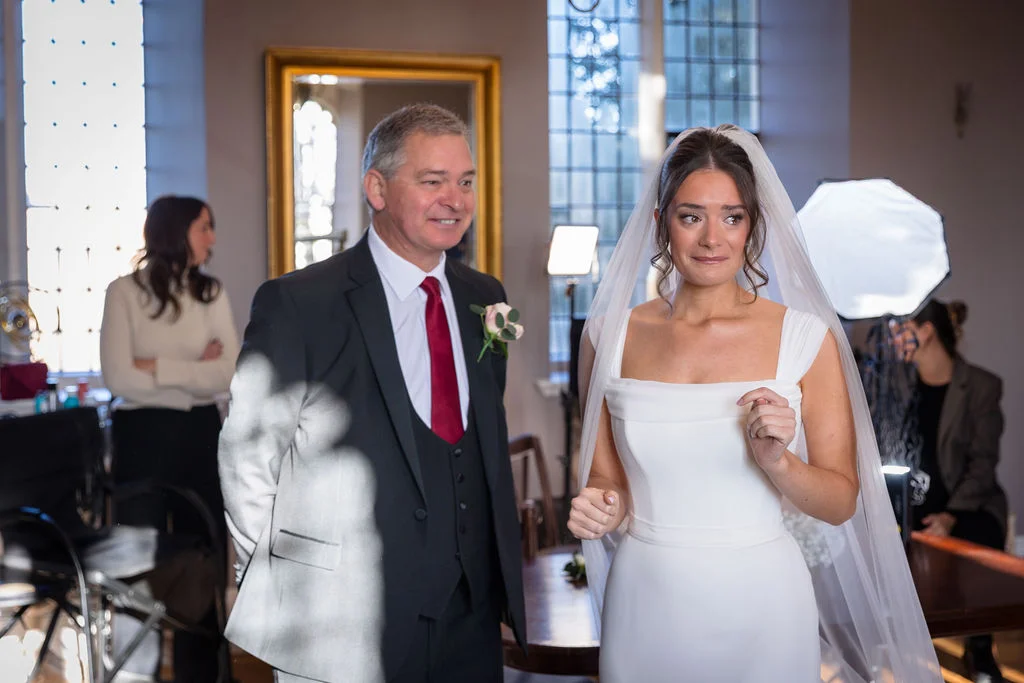 An emotional bride with tears in her eyes stands next to her father in a suit during wedding preparations indoors.