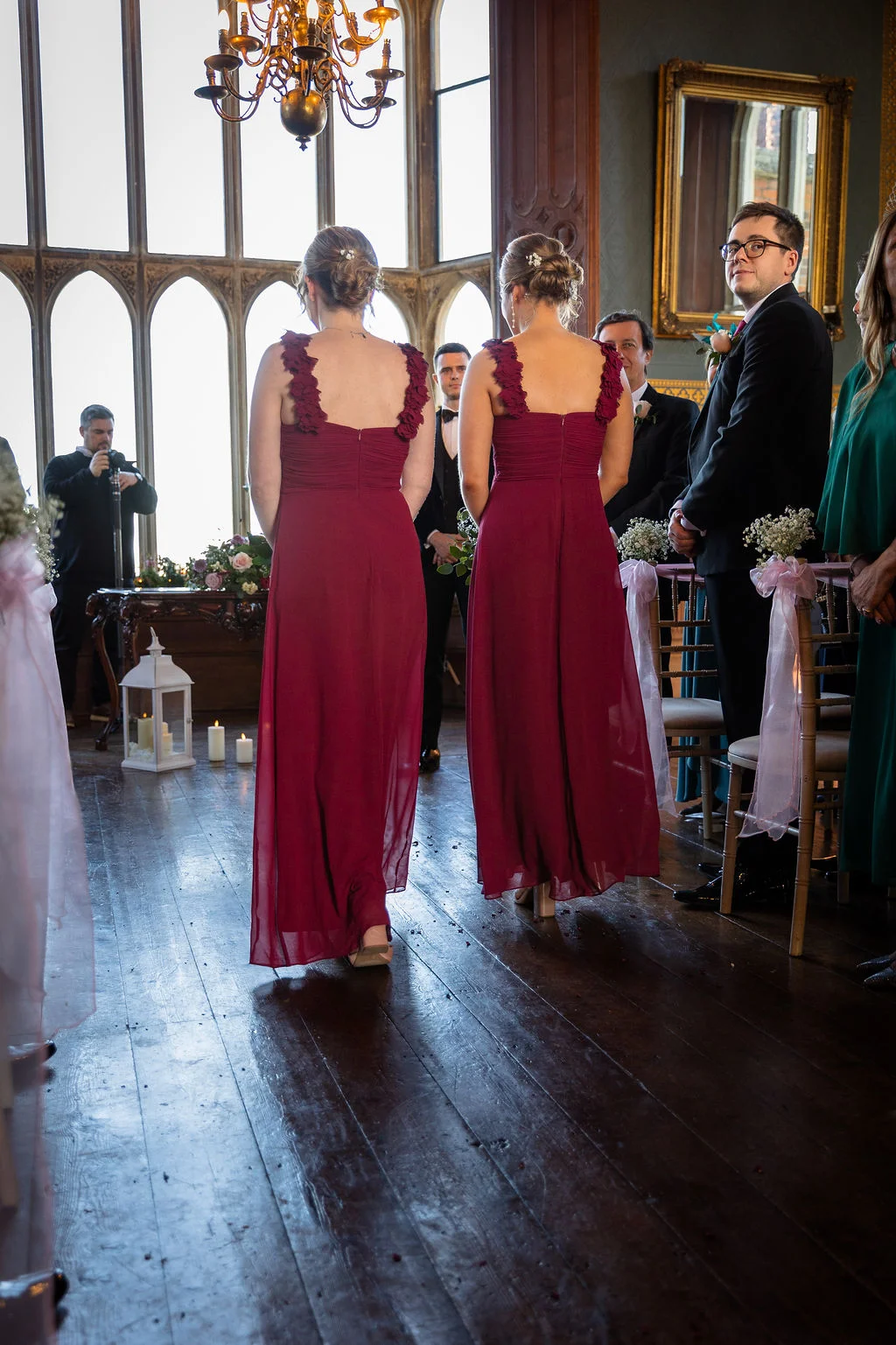 Two bridesmaids in matching burgundy dresses walk down the aisle during a ceremony inside a grand country house venue with large windows and wooden floors.