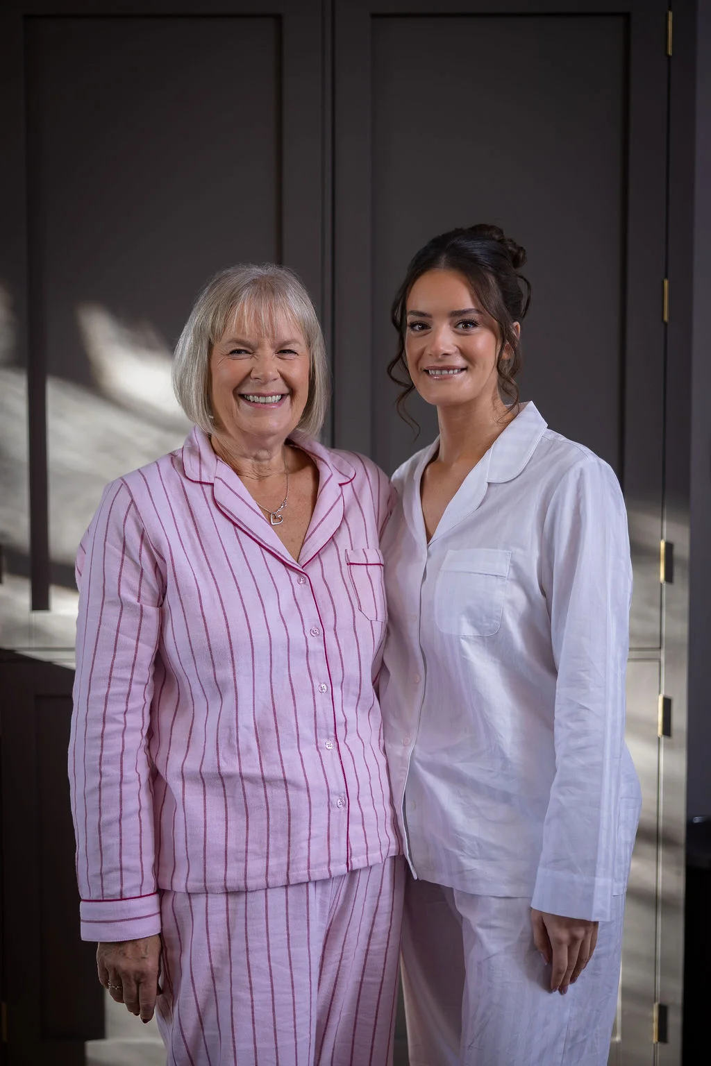 A smiling bride wearing white pyjamas stands side by side with her mother in matching pink-striped pyjamas indoors during wedding morning preparations.