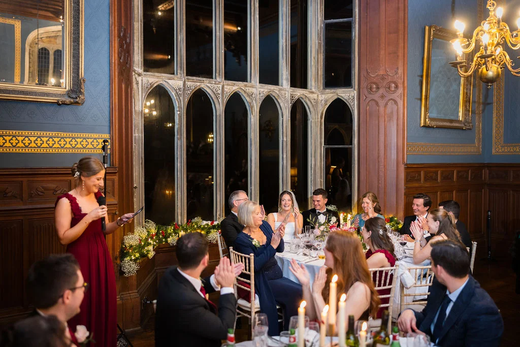 A bridesmaid in a burgundy dress delivers a speech at a candlelit wedding reception in a historic wood-panelled hall with large arched windows.