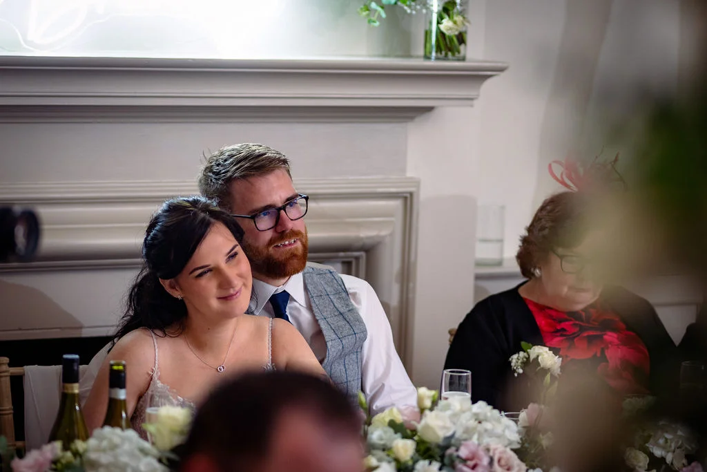 A smiling bride and groom sit at the top table during wedding breakfast speeches, with guests visible nearby in a softly lit room.