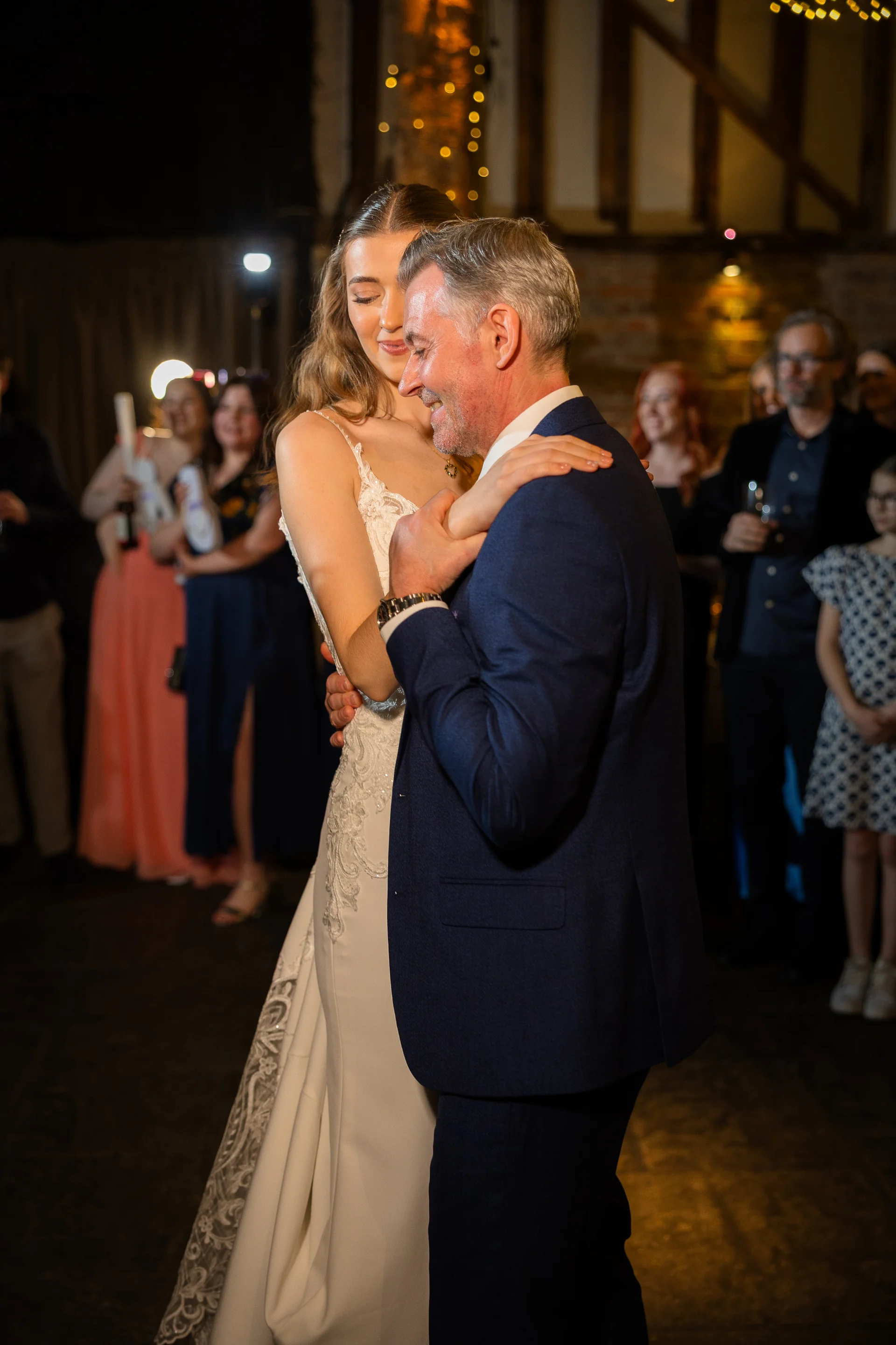 A bride shares an emotional father-daughter dance on the reception floor inside a rustic barn venue with exposed wooden rafters.