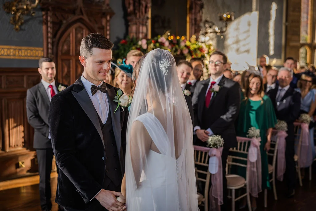 A groom in a black velvet tuxedo looks lovingly at his bride during their church ceremony, surrounded by seated wedding guests.