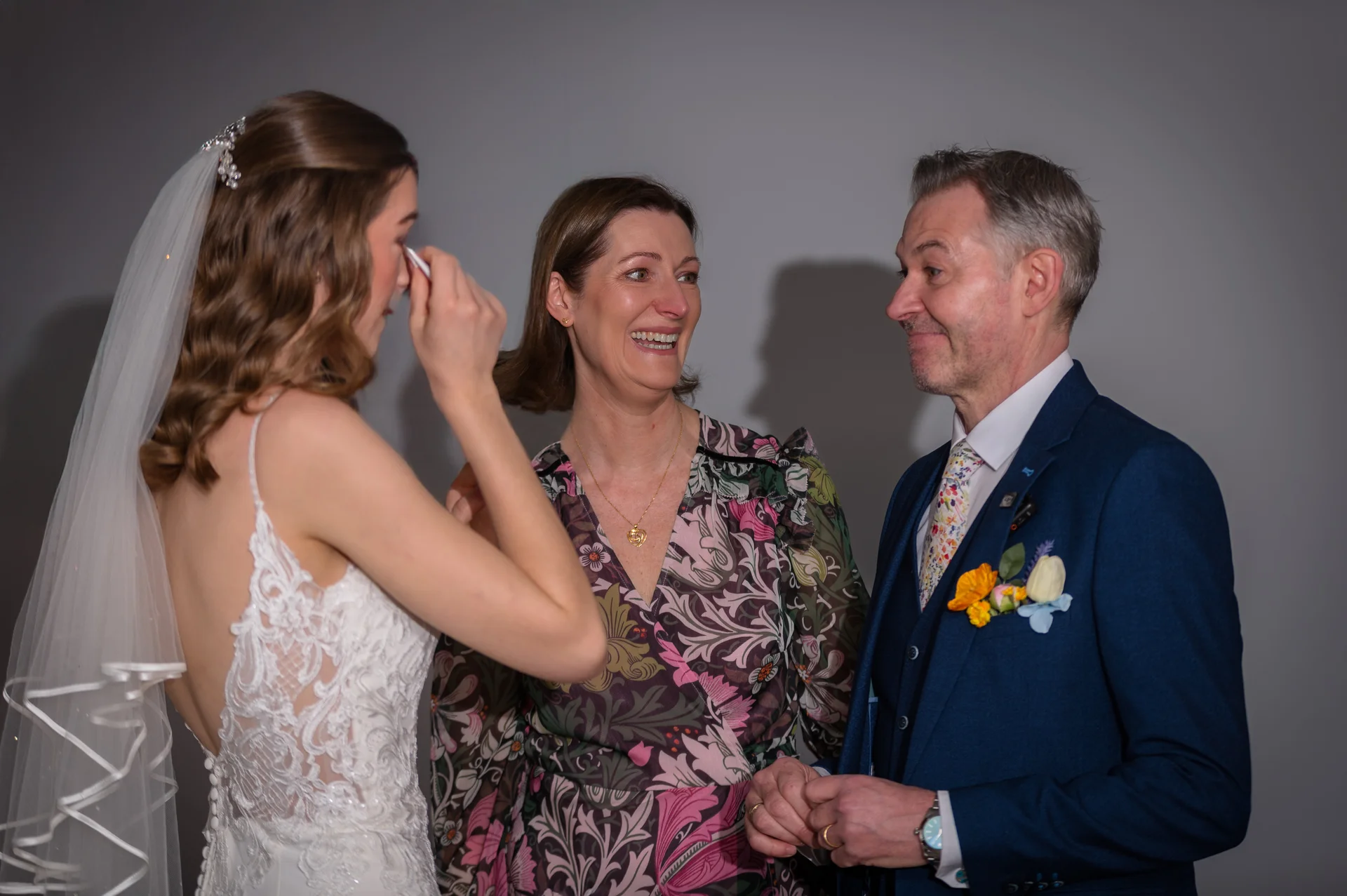 An emotional bride wiping a tear shares a tender moment with her smiling mother and father on her wedding day.