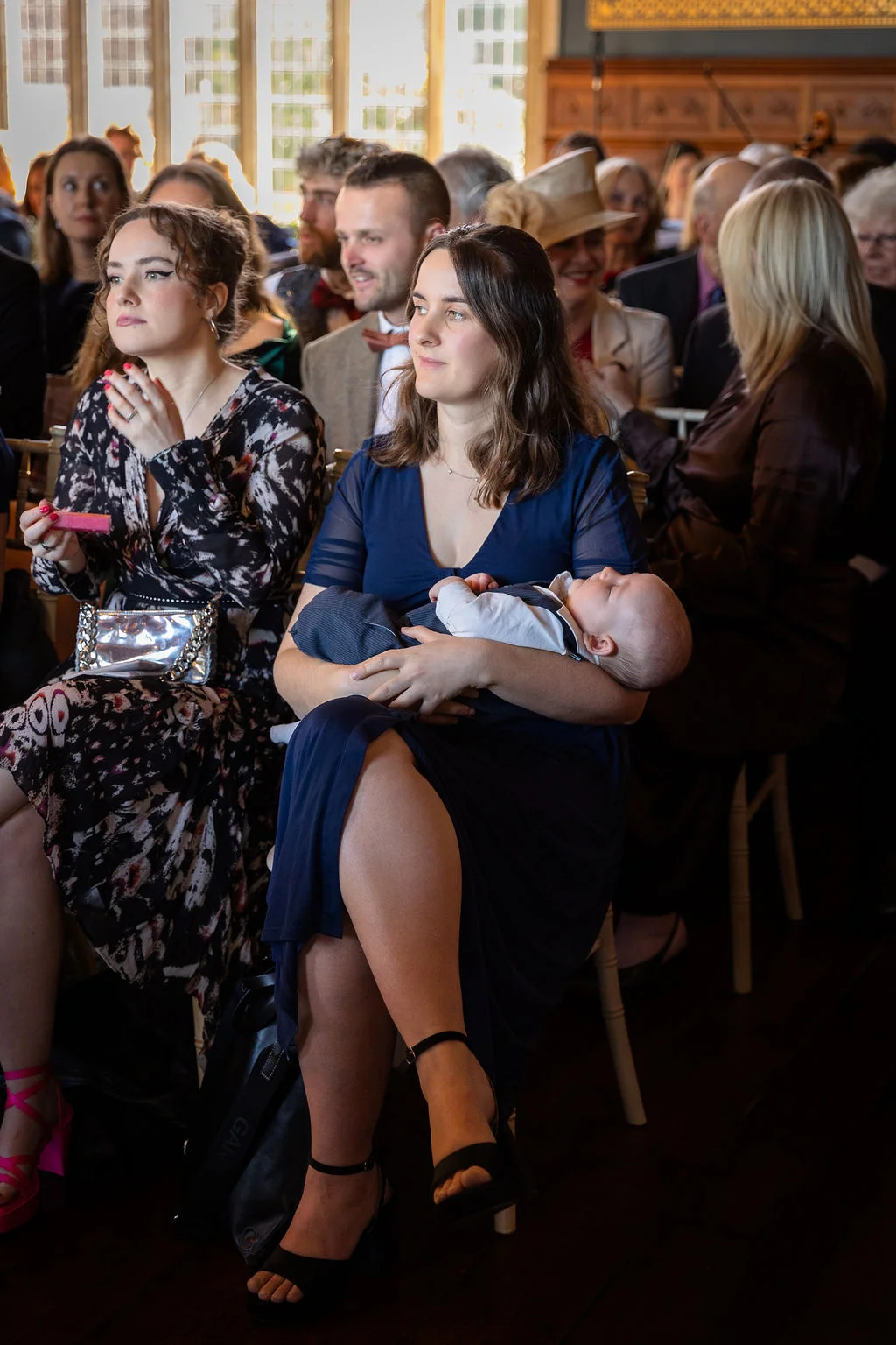 A guest sits amongst a wedding audience holding a sleeping baby on her lap during the ceremony.