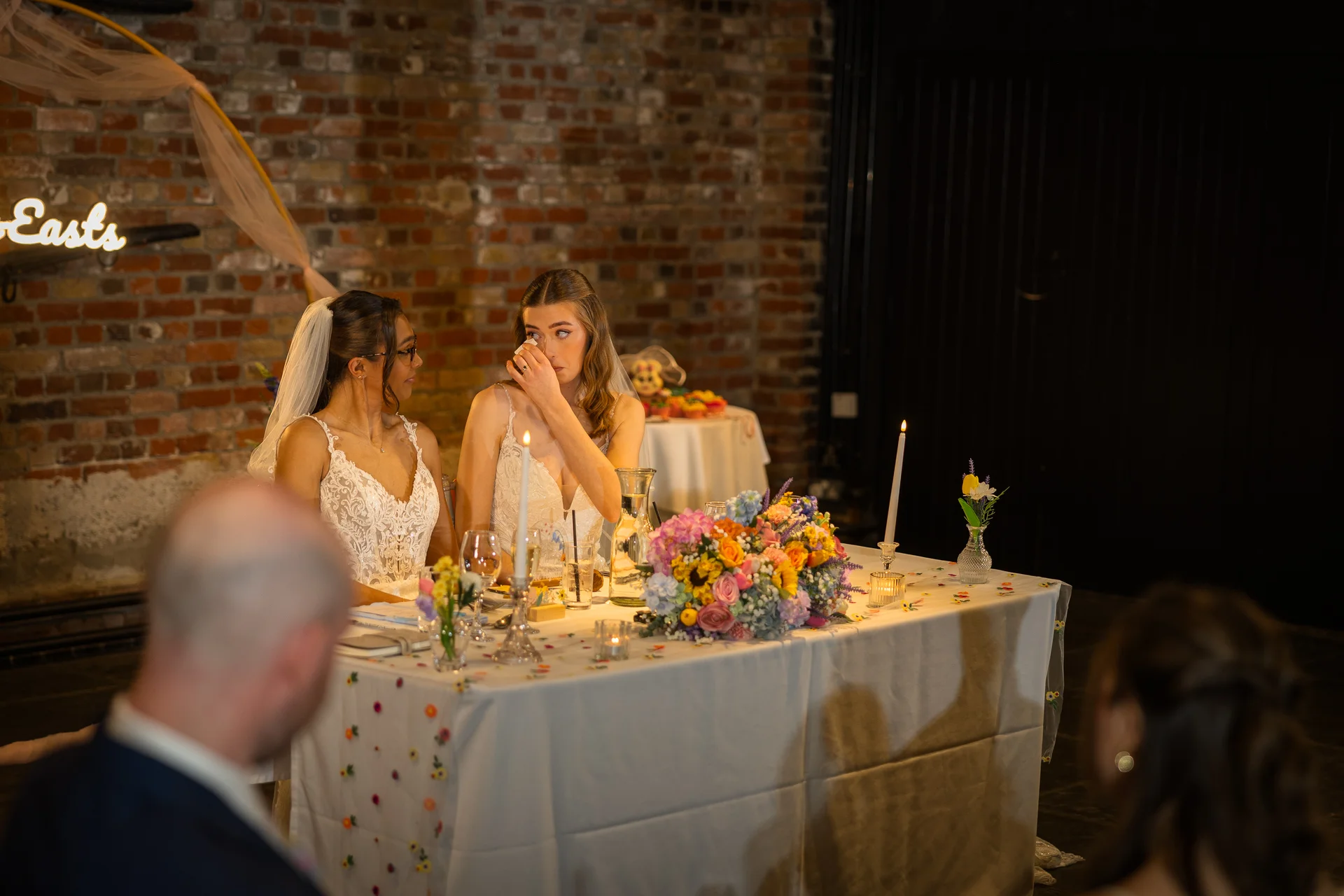 Two brides in lace wedding dresses sit together at a top table in a rustic brick venue, with one bride wiping away tears of emotion during speeches.