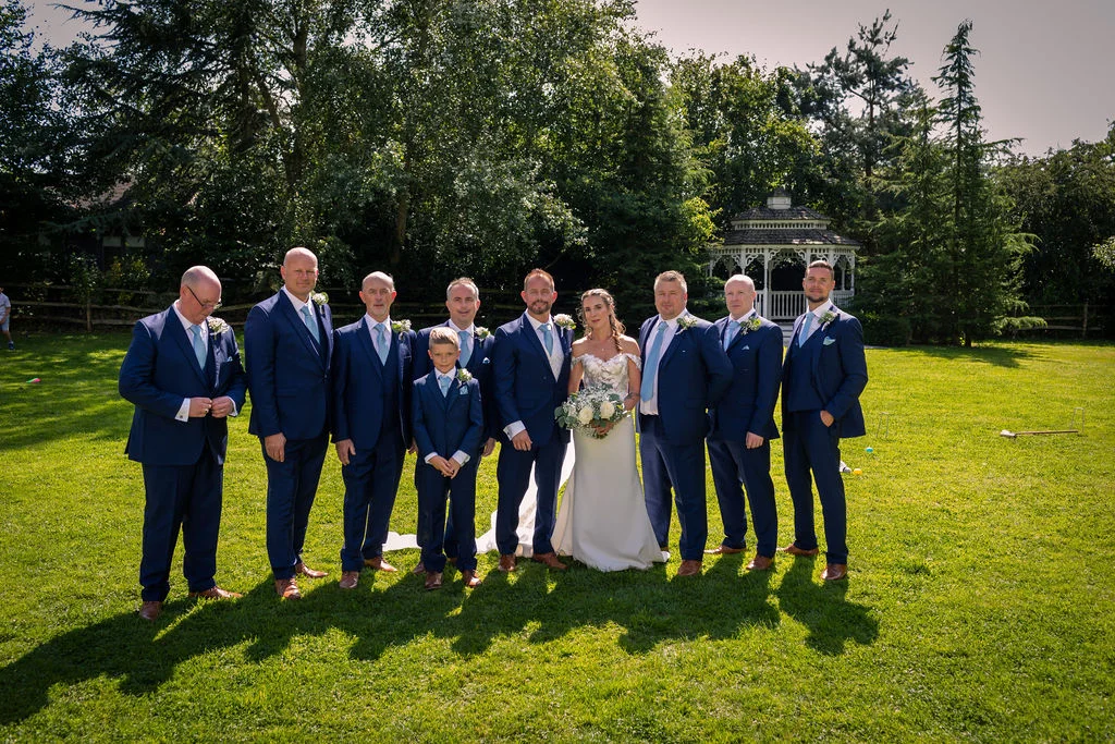 A bridal party poses outdoors on a sunlit lawn in front of a white gazebo, with the bride in a white gown holding a bouquet surrounded by groomsmen in matching navy suits.