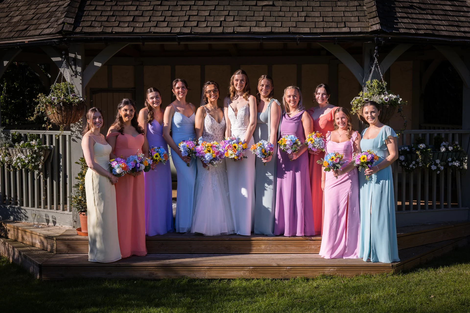 A smiling bride in a lace gown and veil stands proudly at the centre of a large group of mismatched bridesmaids wearing colourful pastel dresses outside a wooden gazebo on a sunny day.