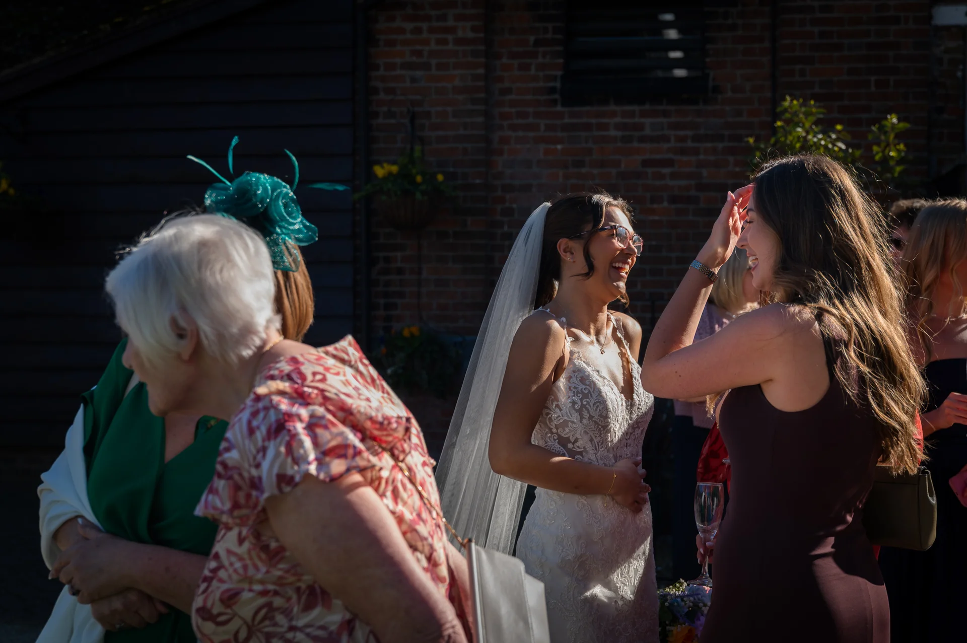 A smiling bride wearing glasses and a lace wedding dress shares a joyful moment with guests during a sunny outdoor drinks reception.