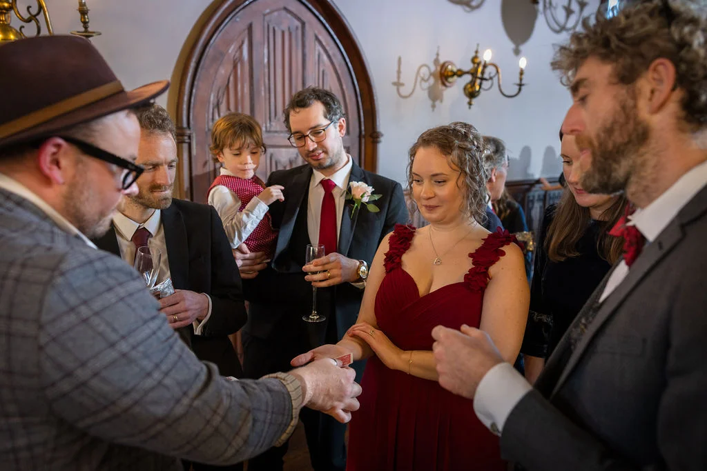 A close-up magic performer shows a card trick to an amused group of wedding guests, including a woman in a deep red gown.