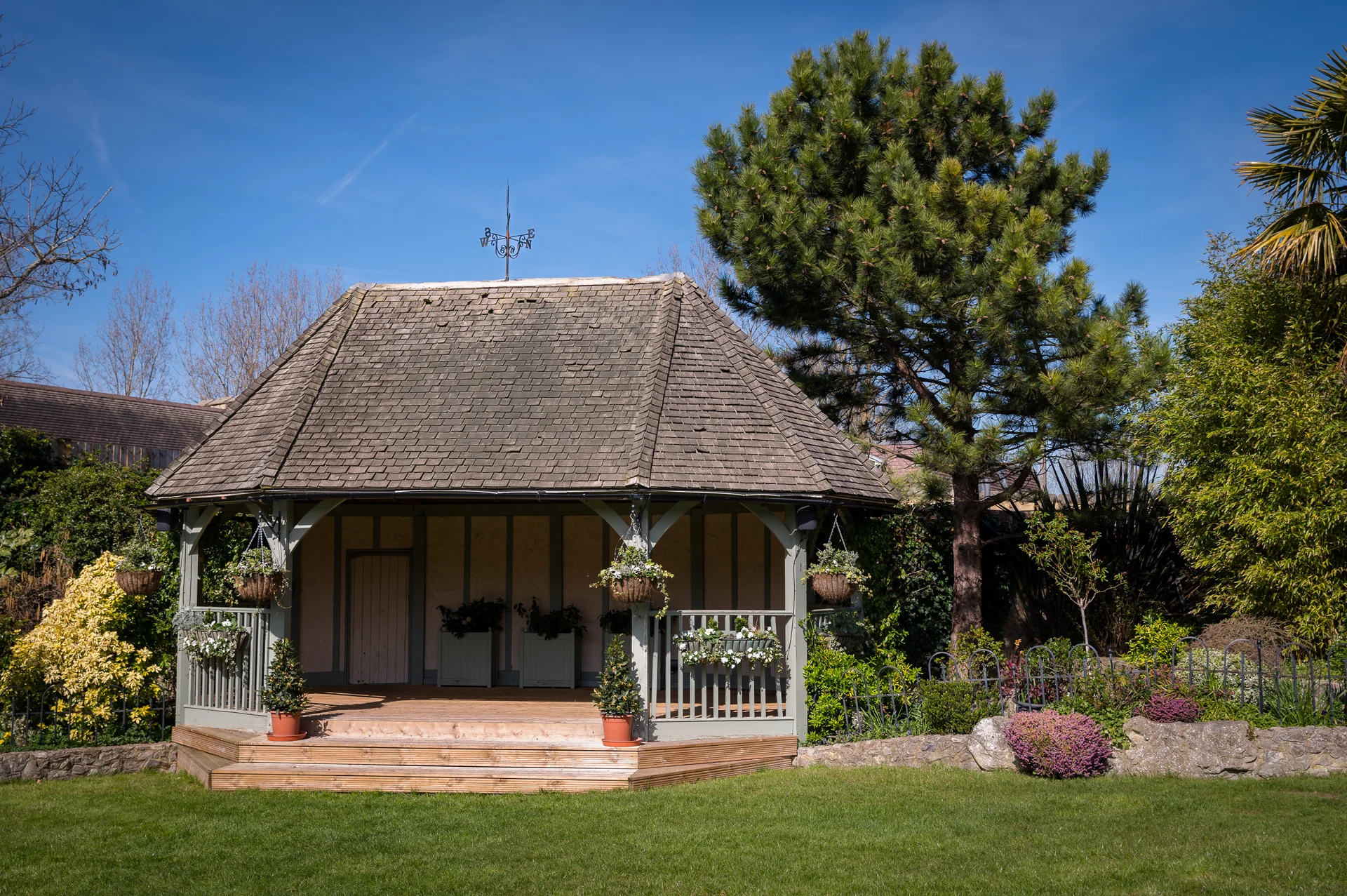A charming wooden garden gazebo stands on a manicured lawn under a bright blue sky, surrounded by lush foliage and blooming flowers.
