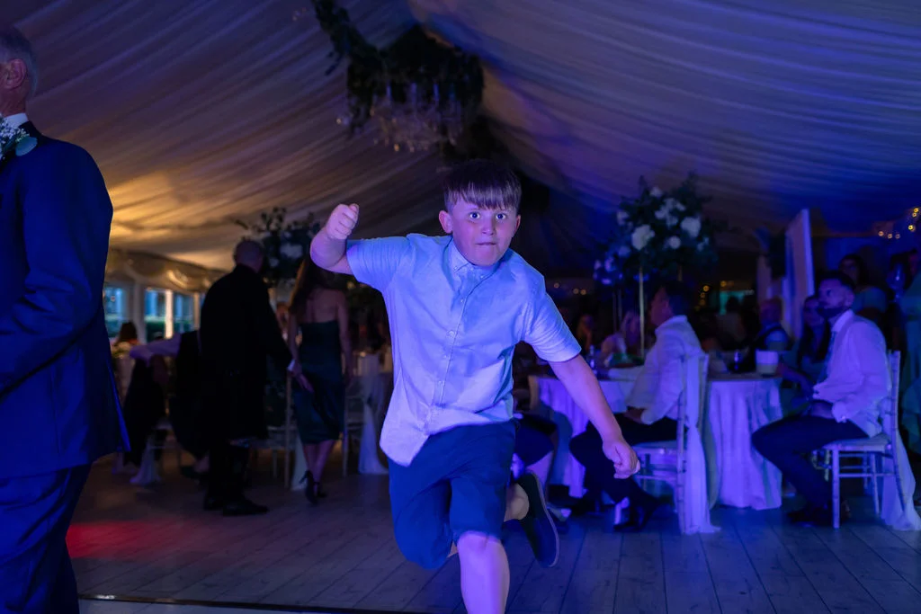 A young boy energetically dances on the wooden floor of a marquee reception bathed in cool blue lighting.