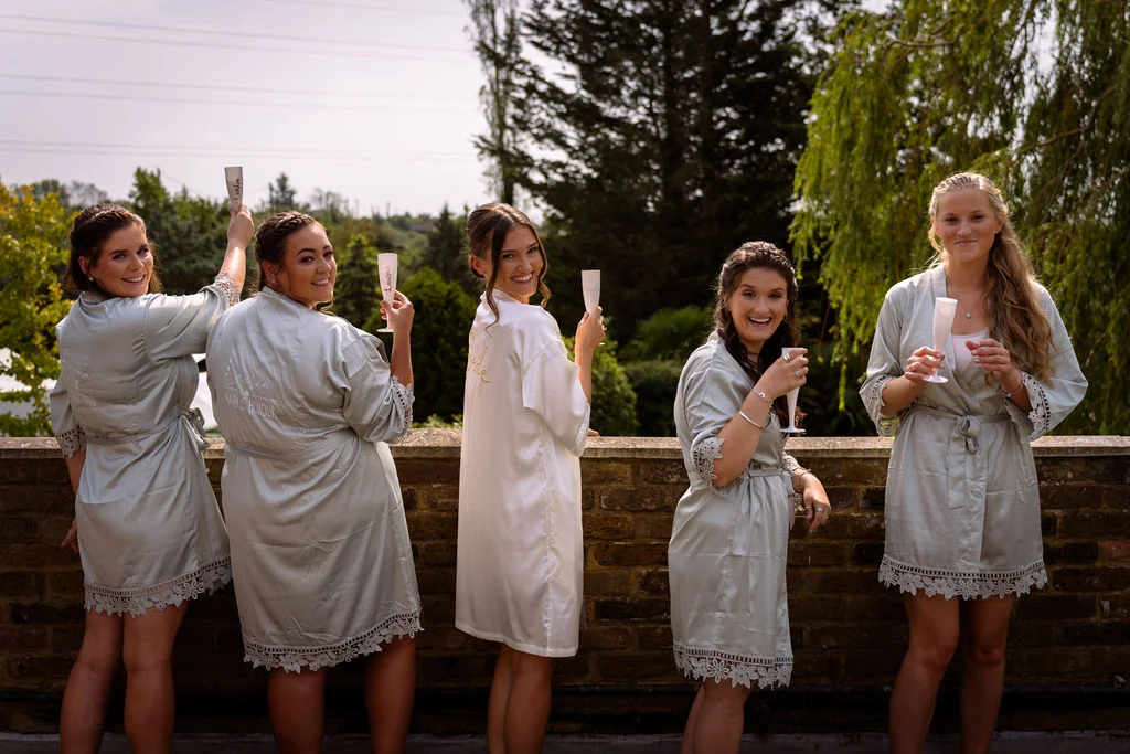 Five bridesmaids and a bride wearing matching satin robes stand against a brick wall, holding champagne glasses and laughing over their shoulders.
