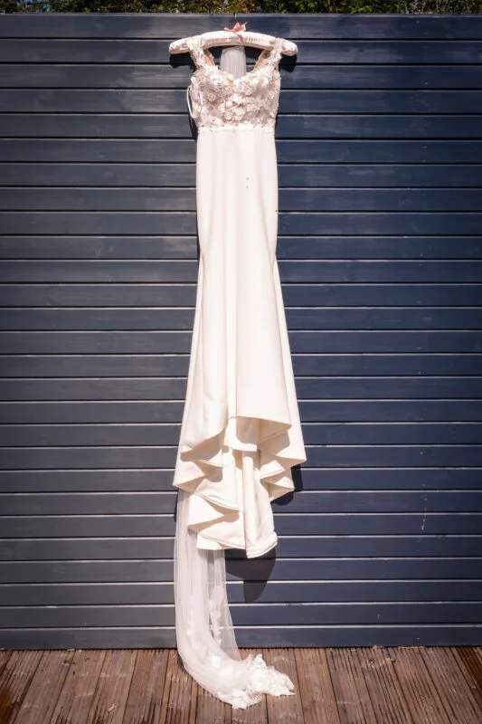 A stunning white crepe wedding gown with a floral lace bodice and a sweeping train hangs against a dark grey slatted wooden wall.