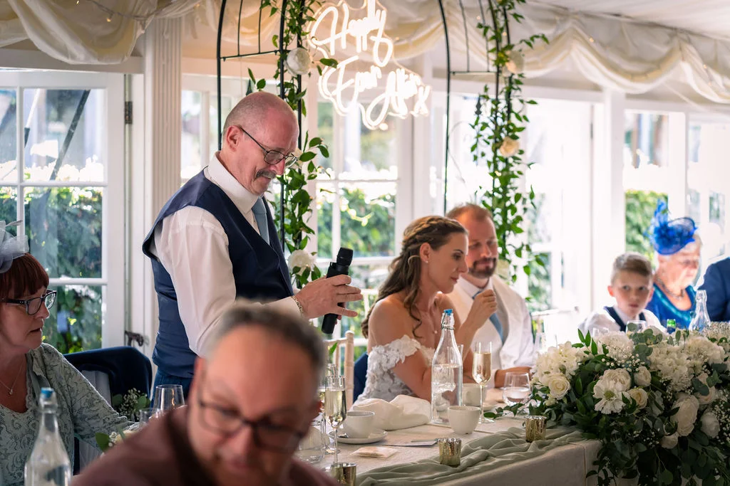 A proud father of the bride delivers a speech at the top table inside a brightly lit marquee decorated with a floral runner and a glowing neon sign.
