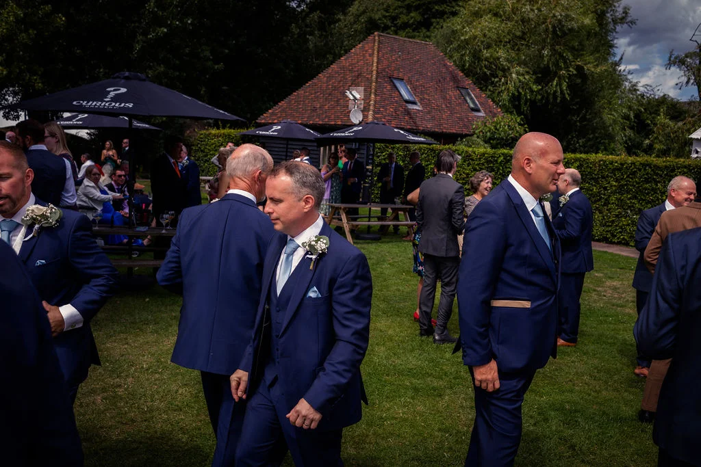 Wedding guests in navy suits and casual attire mingle on a lush green lawn during an outdoor afternoon reception.