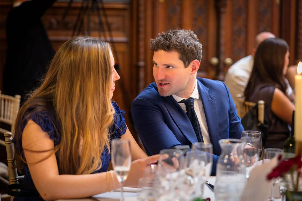 A guest in a navy suit leans in to converse with a female guest seated at a wedding breakfast table in a wood-panelled reception room.