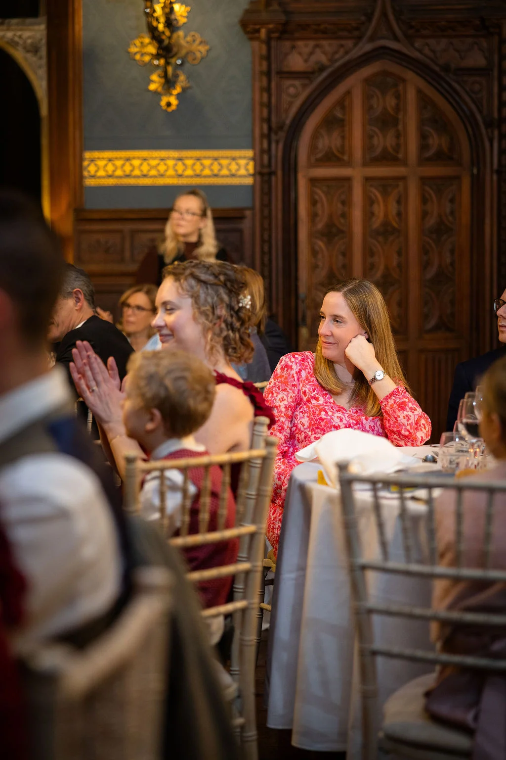 Wedding guests gathered around round tables listen attentively during the reception speeches inside a historic wood-paneled hall.