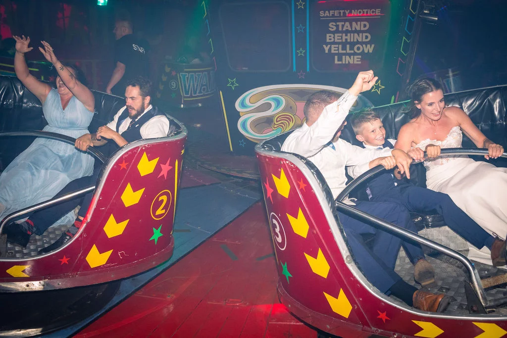 Wedding guests and a bride in her gown enjoy a funfair ride together, laughing and raising their hands in the air with direct flash photography.