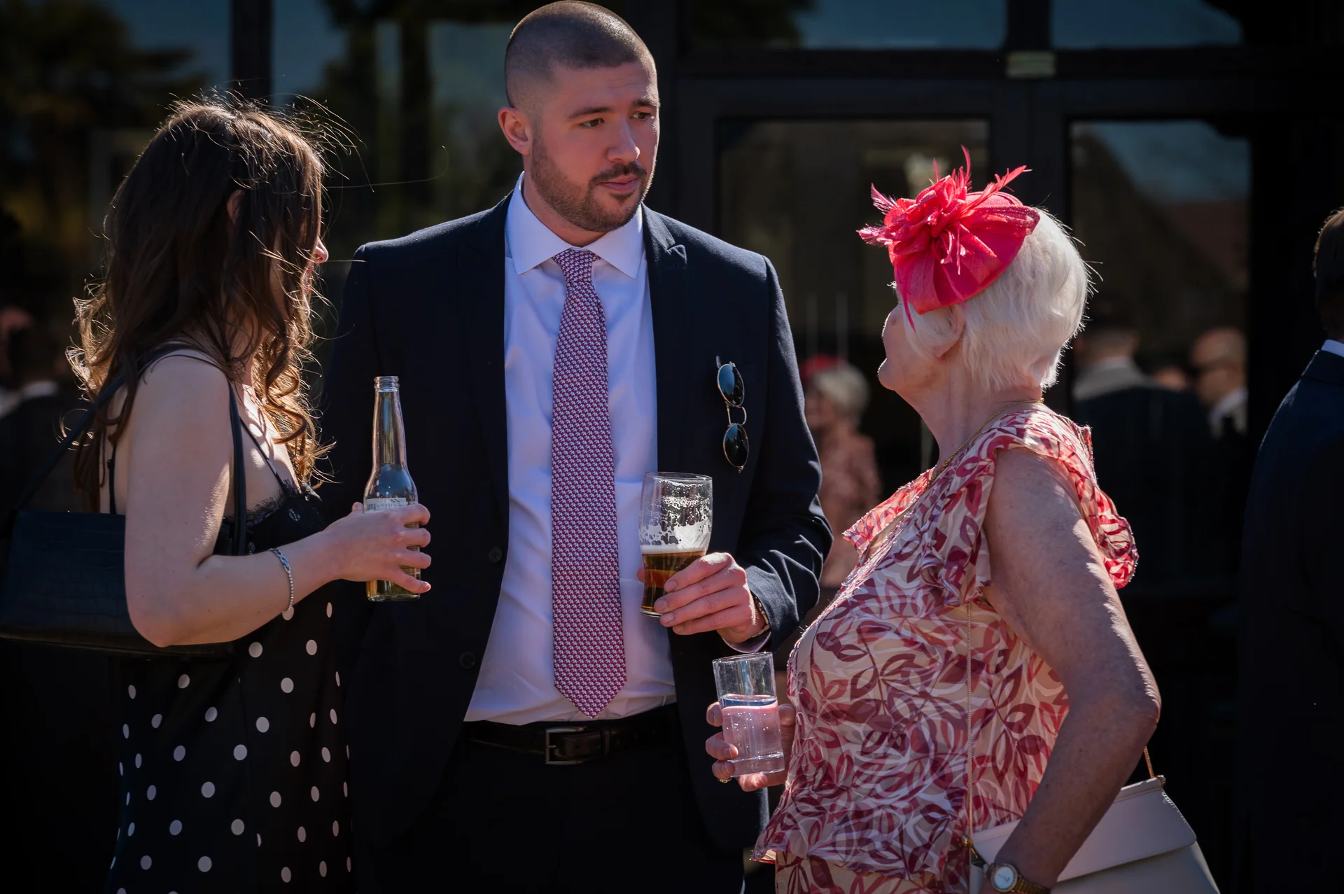 Wedding guests mingle and converse outdoors during a sunny drinks reception, holding drinks and wearing smart attire and a vibrant fascinator.