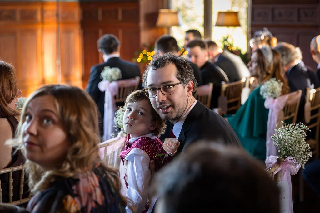 A smiling man holding a young page boy in a red patterned vest during a wedding ceremony in a wood-panelled room.