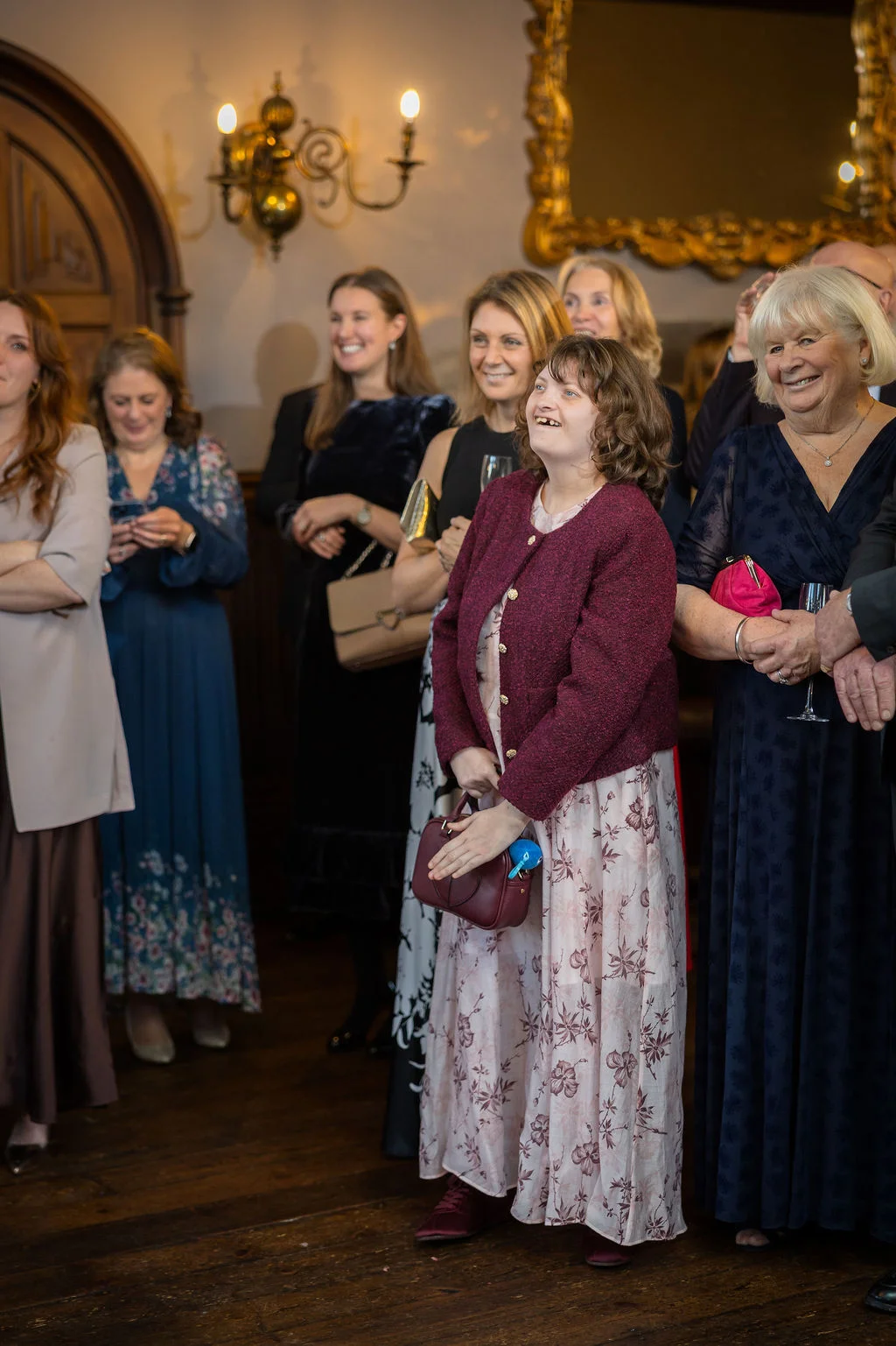 A group of smiling wedding guests gather indoors, watching the reception speeches with joy and amusement.