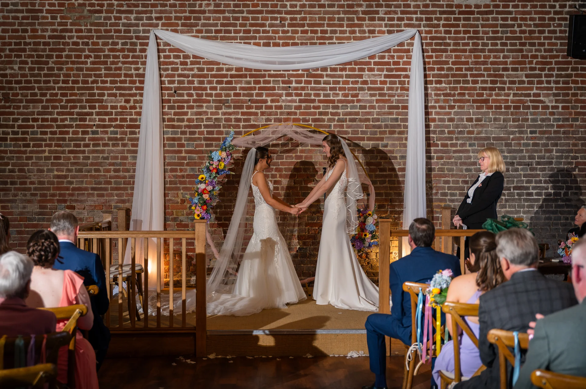 Two brides hold hands during their wedding ceremony in front of an exposed brick wall decorated with a fabric backdrop and floral arch.