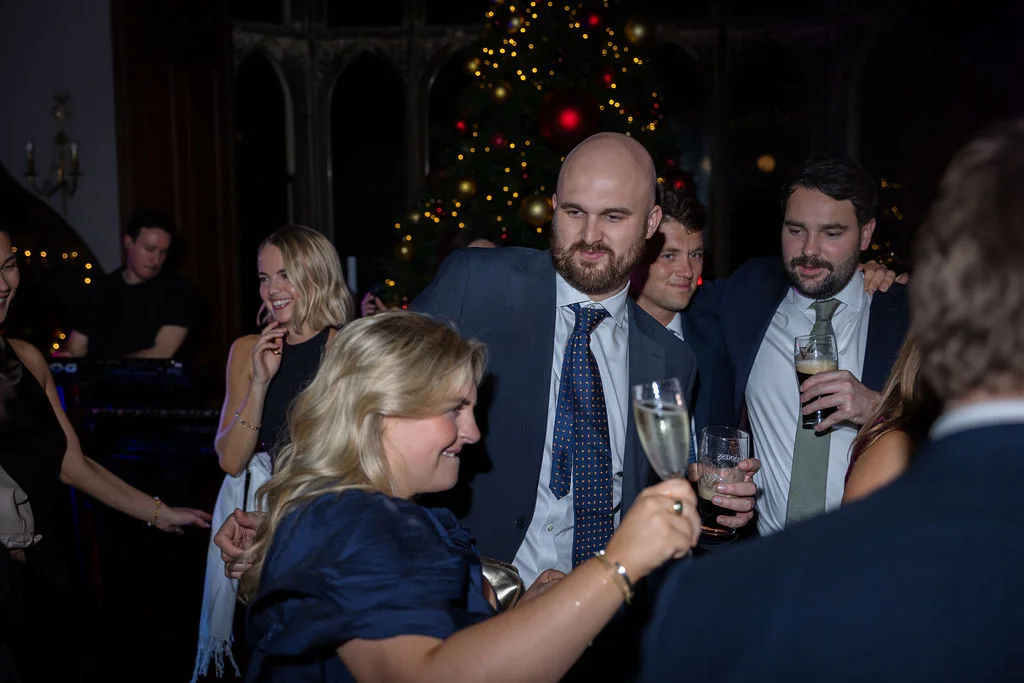 Wedding guests enjoy drinks and laughter on the dance floor during a festive evening reception illuminated by a softly glowing Christmas tree.