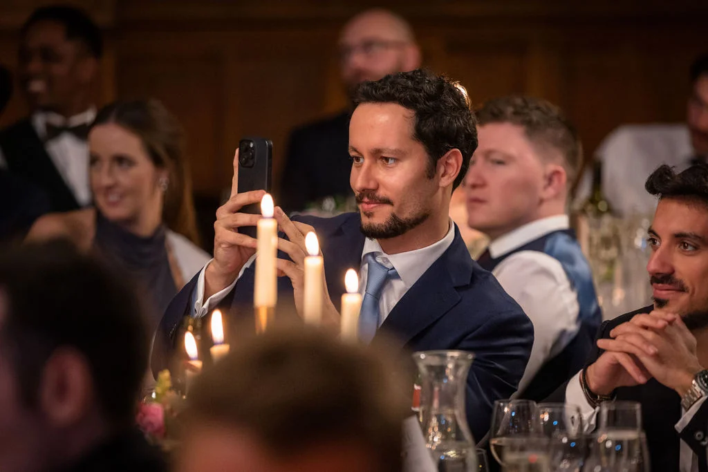 A guest in a navy suit uses his smartphone to capture a moment during a candlelit wedding reception surrounded by other guests.