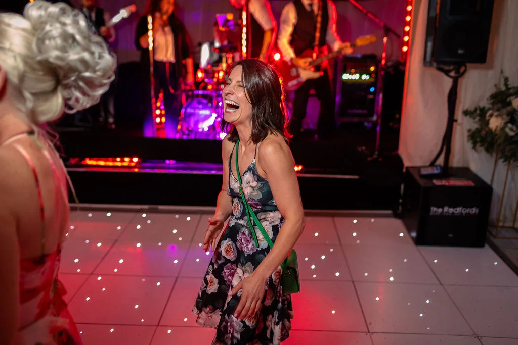 A wedding guest wearing a floral dress laughs joyfully while dancing on an illuminated dance floor as a live band performs on stage behind her.
