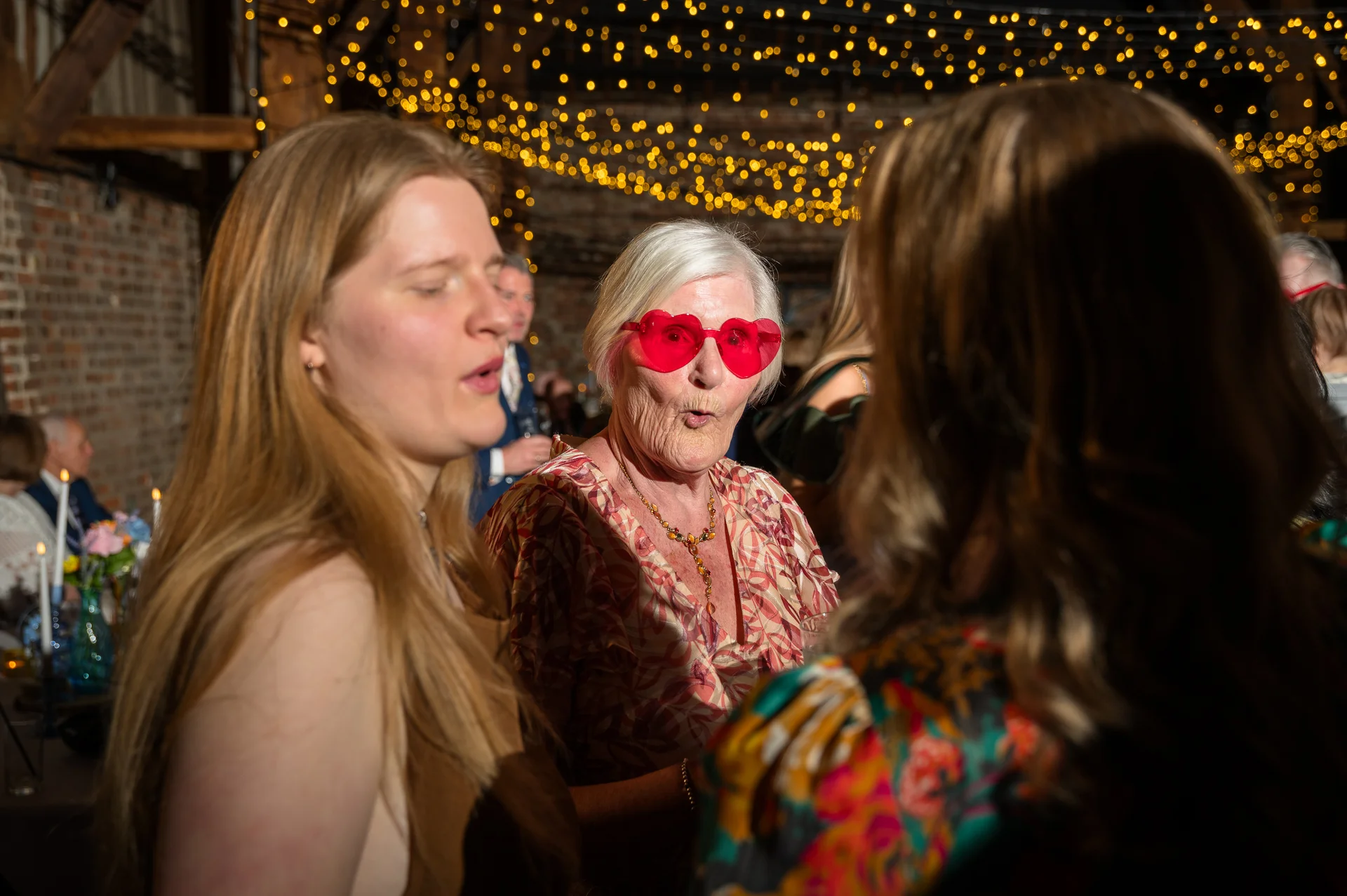 An elderly female wedding guest wearing playful heart-shaped glasses smiles amidst a lively evening reception illuminated by a canopy of fairy lights.